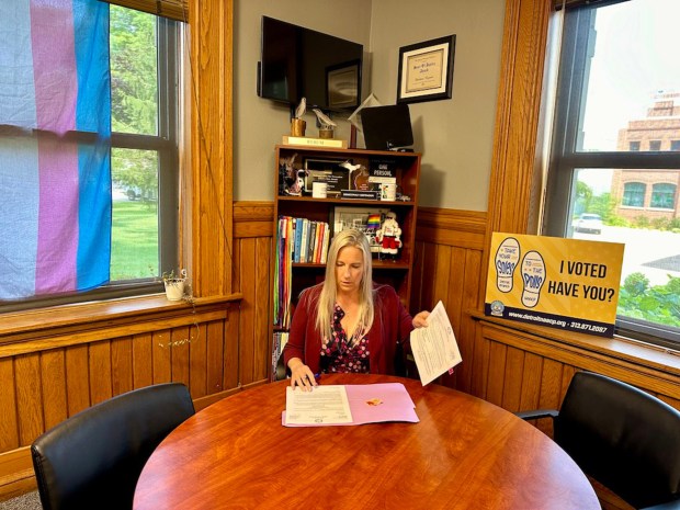 A woman sits at a table in her home flanked by a trans rights flag and an "I voted" sign